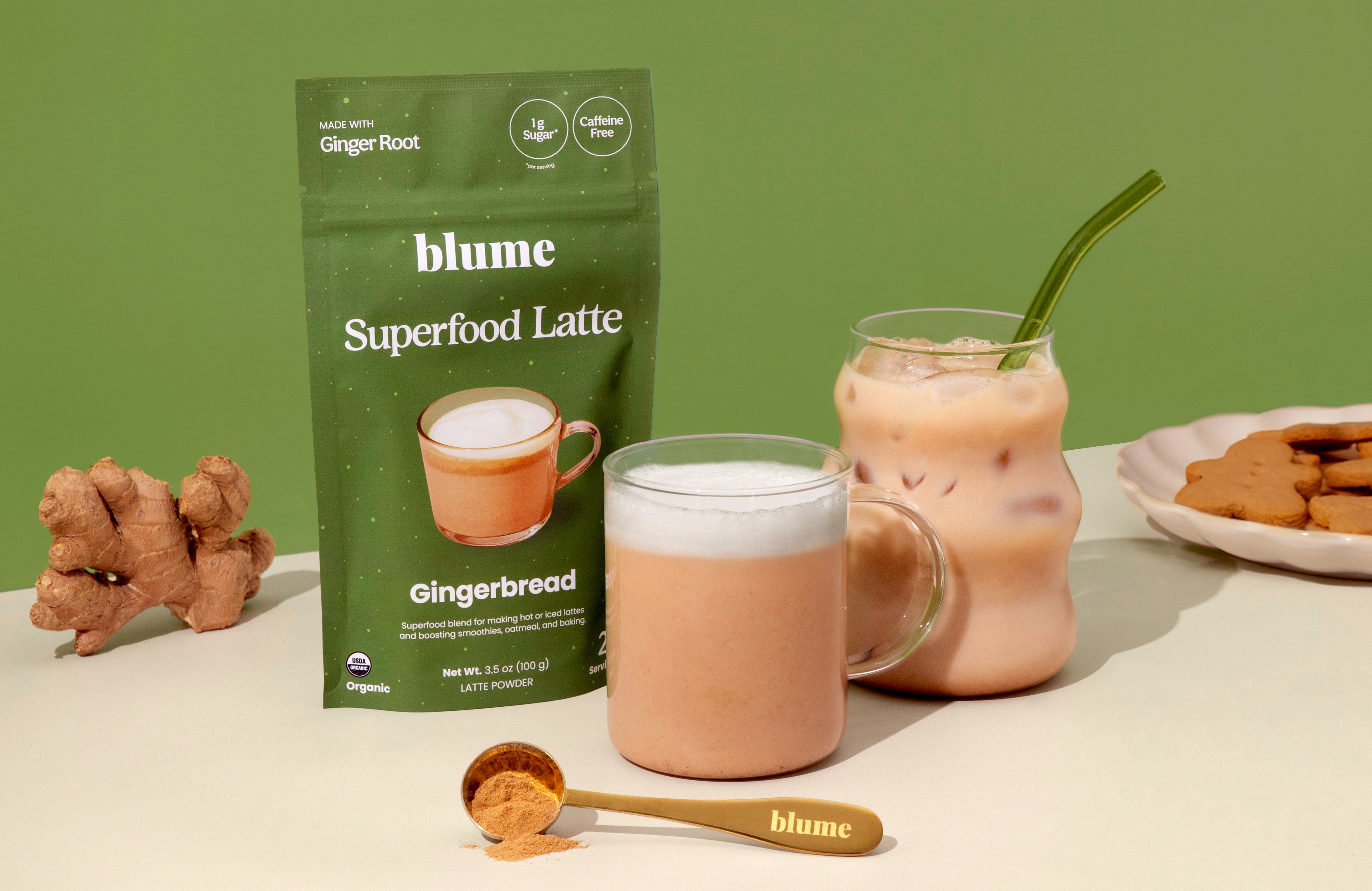 A green packet of gingerbread superfood latte powder is next to a glass of latte and an iced latte. A spoon with powder, fresh ginger, and a plate of gingerbread cookies are nearby, all set against a green background.